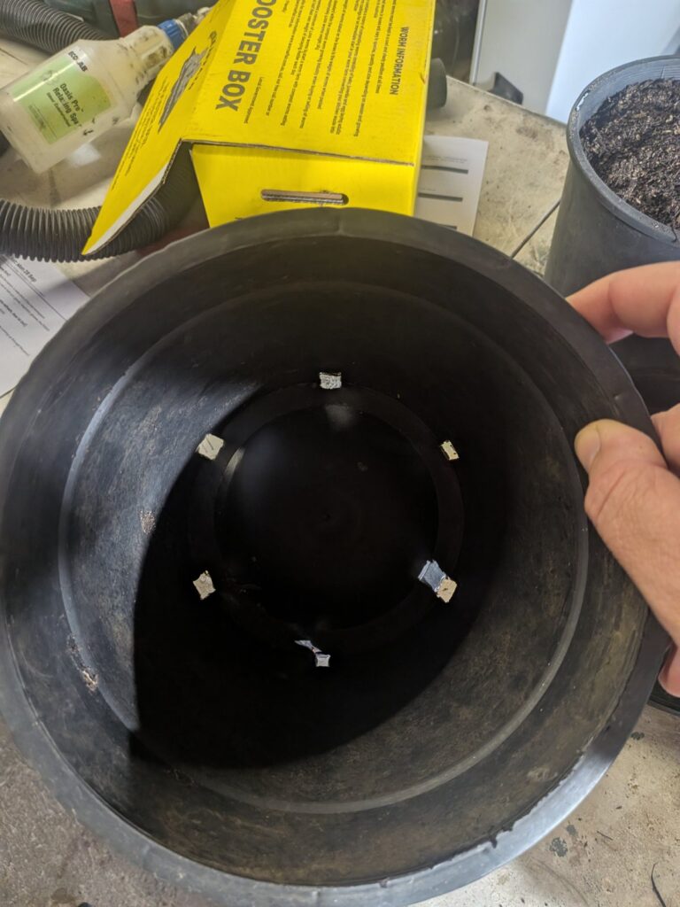 Black nursery pot on a saucer next to a 25 L bag of all-purpose potting mix, a hand trowel and a packet of mixed salad leaf lettuce seed